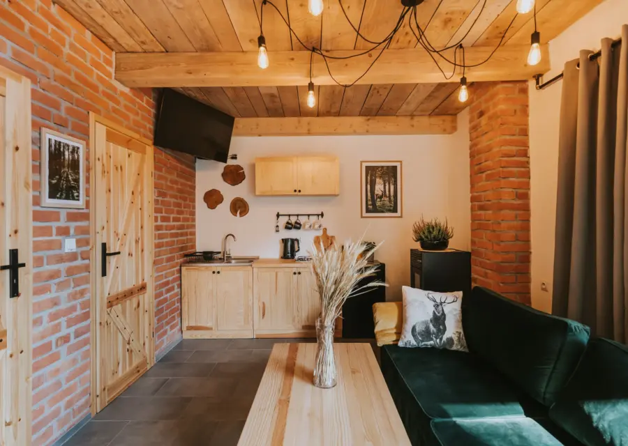 Salon z aneksem kuchennym w domku Jeleni Szlak: ceglana ściana, drewniany stół i zielona sofa